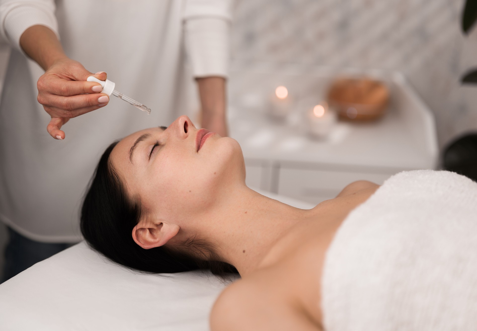 Young lady lying with closed eyes while cosmetician applying essential oil on face